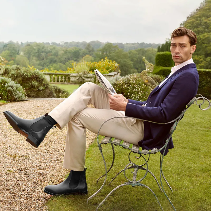 A stylish man in a navy blazer and light trousers sits on a metal garden chair reading a newspaper, wearing sleek black leather platform Chelsea boots.